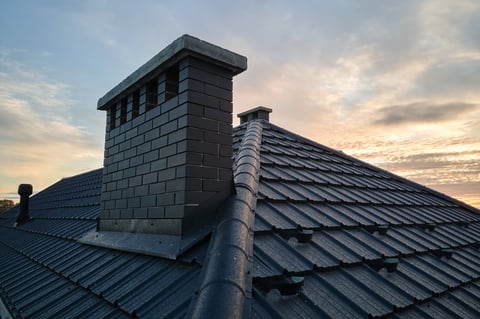 A dark metal roof with an ornate brick chimney against a cloudy sunset sky