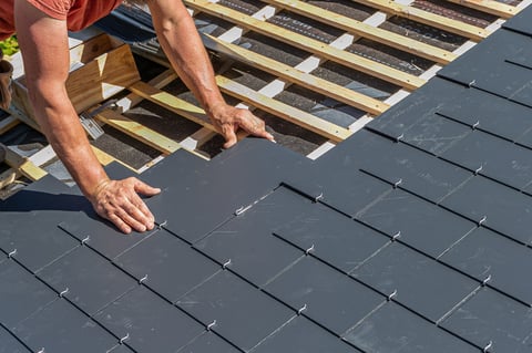 Worker installing dark roof tiles on wooden roof frame during construction or renovation