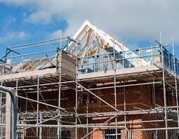 House under construction with metal scaffolding, exposed wooden framing, and white roof trusses against blue sky