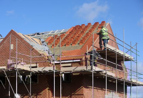 Brick house under construction with metal scaffolding, exposed roof framing, and red tiles against blue sky